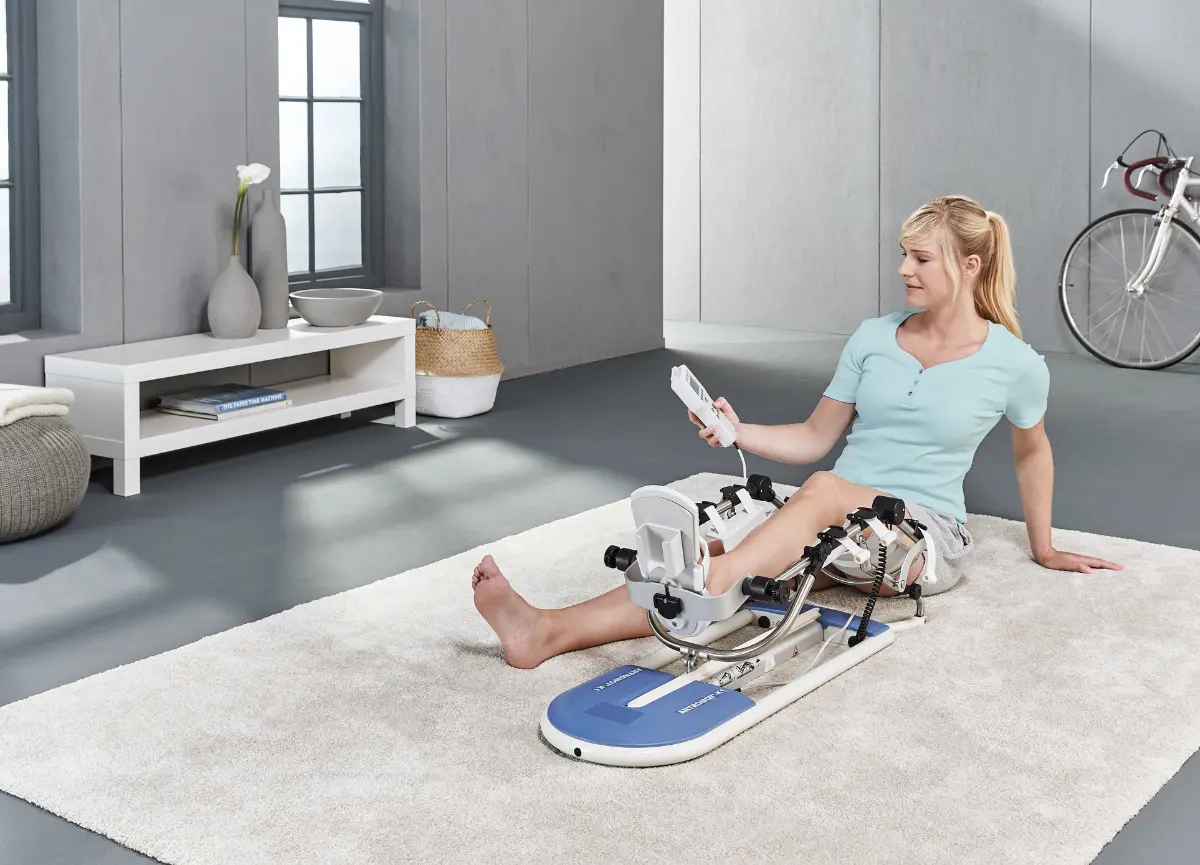 A woman using a CPM (Continuous Passive Motion) machine, the Artromot K1, while sitting on a rug in a modern living room. The individual is holding a remote control and has one leg positioned in the machine for knee rehabilitation.