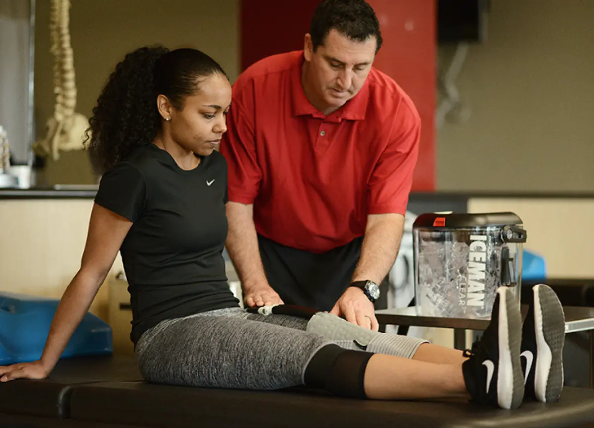 A healthcare professional applying a DonJoy Iceman Clear 3 cold therapy unit to a patient's knee, with the patient seated on a therapy table, wearing athletic clothing, and the Iceman device visible in the background