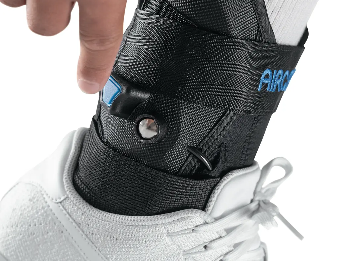 Close-up view of a person adjusting the AirLift PTTD (Posterior Tibial Tendon Dysfunction) brace on their foot, featuring a secure black strap and a blue adjustment button, worn over a white sock and paired with a white sneaker.